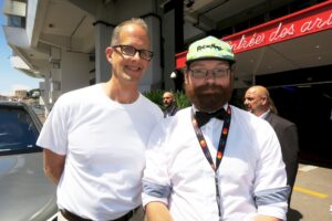 matej-patljak-na-festivalu Matej je na desni strani. Obežen v belo srajco z dolgimi rokavi, ima živo zeleno kapo, okoli vratu ima obesek za ključe, ima očala in brado. Na levi strani je gospod v beli majci in nosi očala.