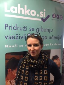 veronika-sustar-13-2-2025 Veronika stoji pred plakatom. Ima spete rjave lase, na glavi ima očala, oblečena v črno oprijeto majco in okoli vratu ima ovit črn šal z rjavimi vzorci, ki predstavljajo črko G.