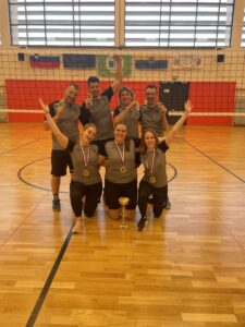 Odbojkarska skupina 7 članov z medaljami okoli vratu. Z nasmehom in z rokami v zraku so postavljeni, da 3 dekleta so na kolenih, zadaj za njimi pa stojijo 3 fantje in 1 dekle.