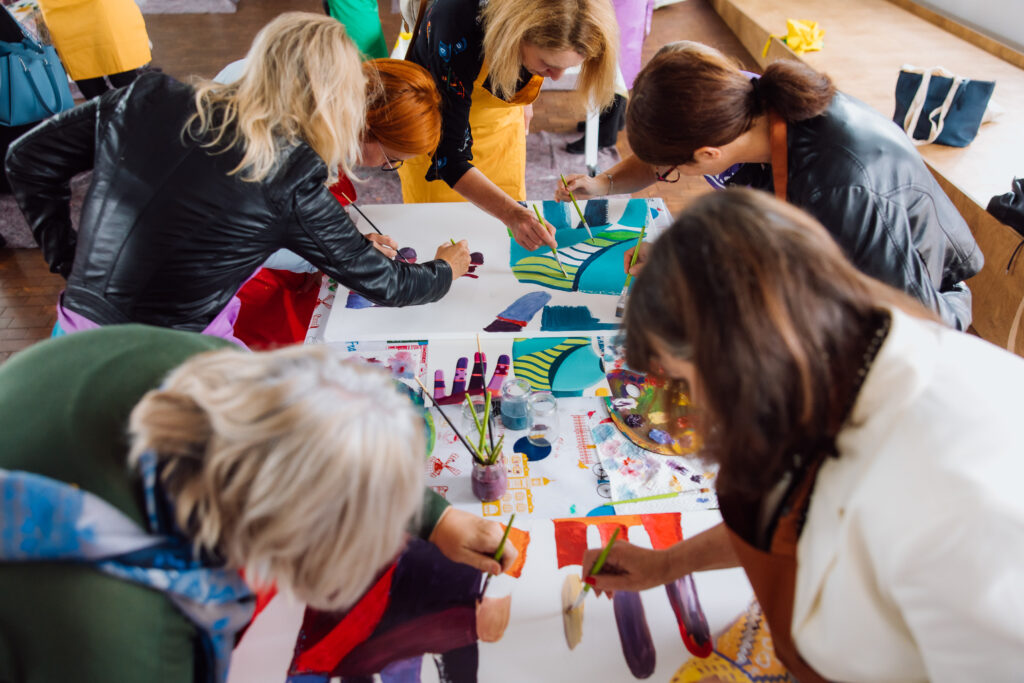 Skupina odraslih žensk (+65) riše s temperami na velik plakat.