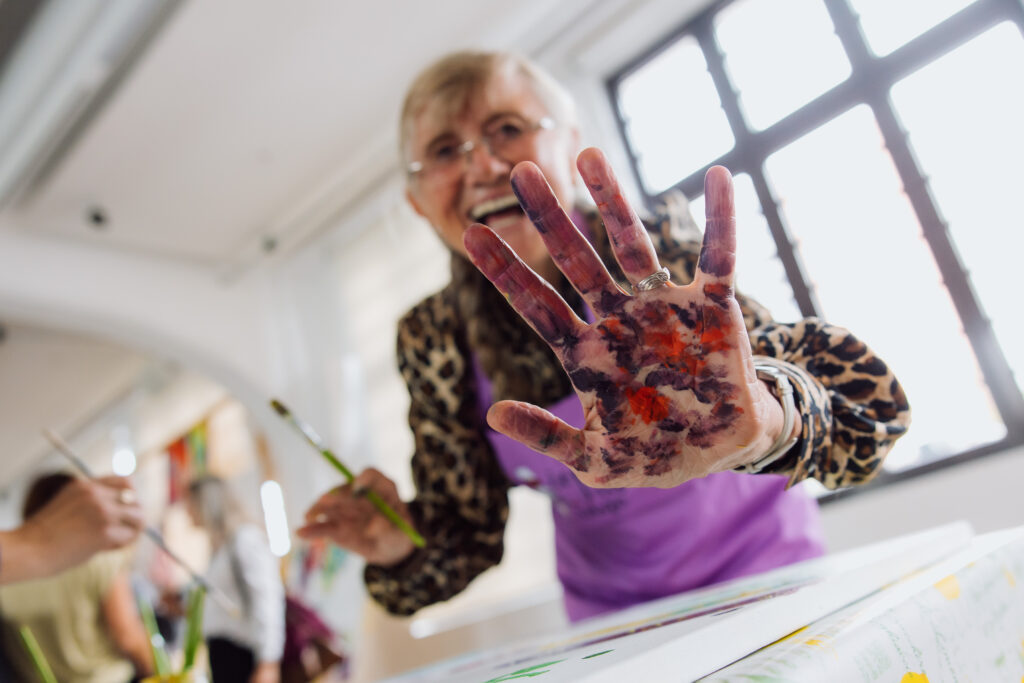 Starejša ženska z nasmehom, nosi očala in lase ima spete, ima iztegnejno roko in kaže dlan, ki je pobarvana s temperami. v drugi roki ima čopič.